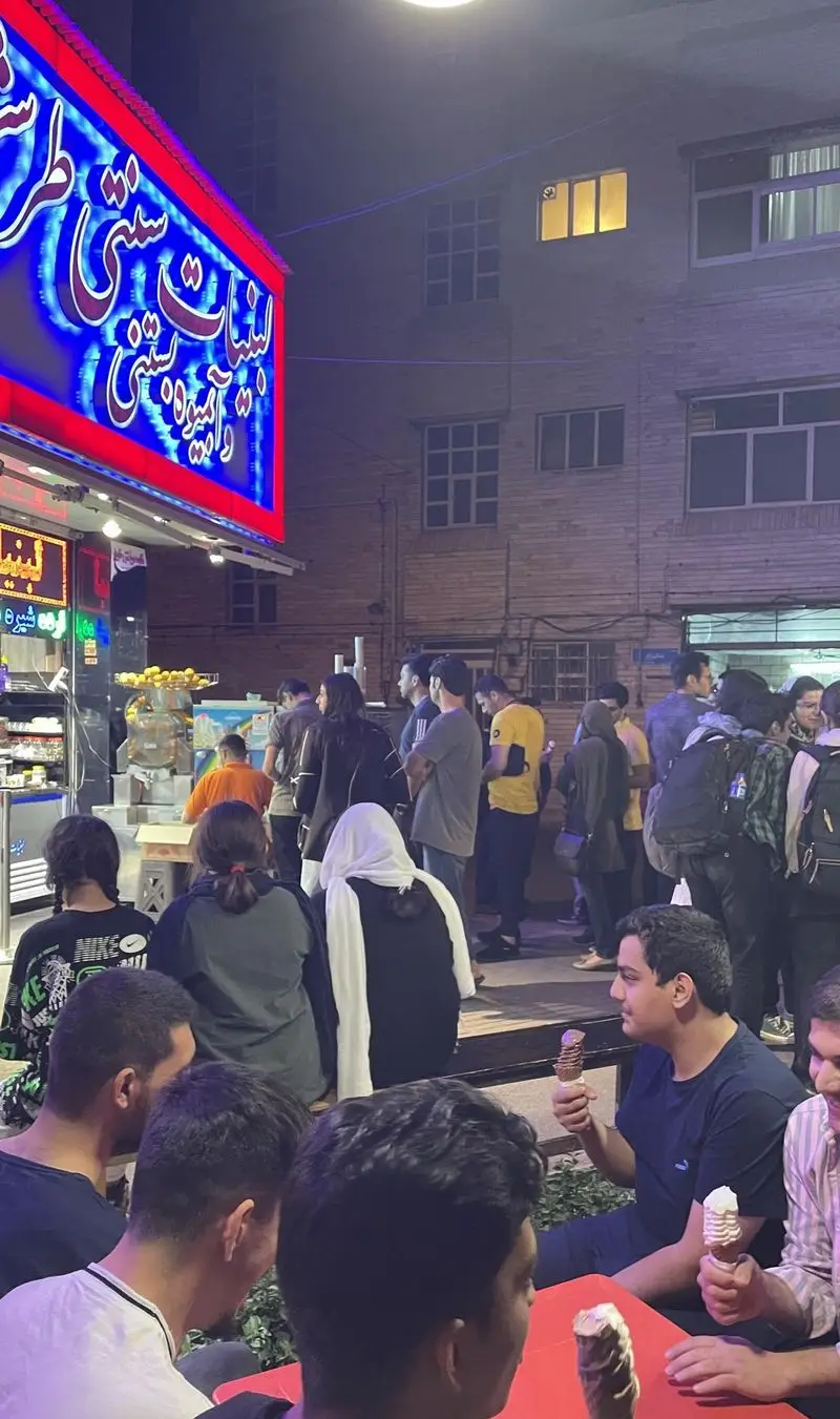 تصویر شبانه از لبنیات سنتی و آبمیوه بستنی طرشت، مملو از مشتریان که از انواع بستنی و آبمیوه تازه لذت می برند.