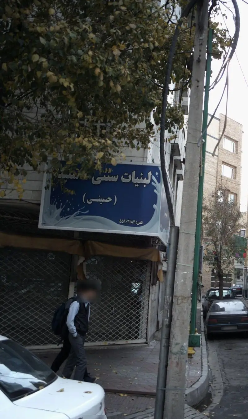 تابلوی مغازه "لبنیات سنتی حسینی" با لوگوی شیر و شماره تلفن ۵۵۹-۲۱۸۲ در نمایی از خیابان شهری و درختان پاییزی.