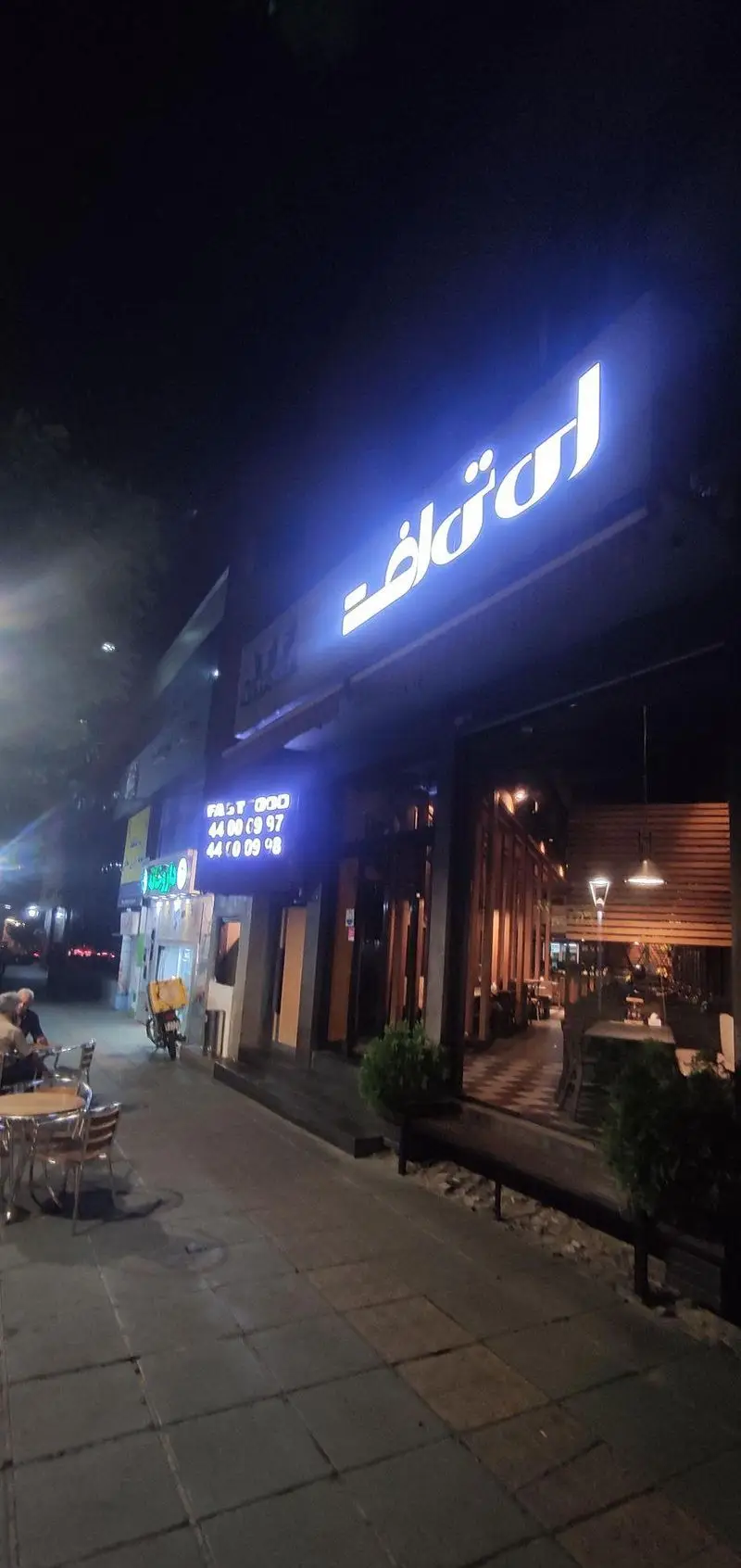 نمای شبانه ای تی اف شعبه کاشانی، فست فود و رستوران با تابلو نئونی روشن، میز و صندلی در فضای باز و موتورسیکلت تحویل غذا.