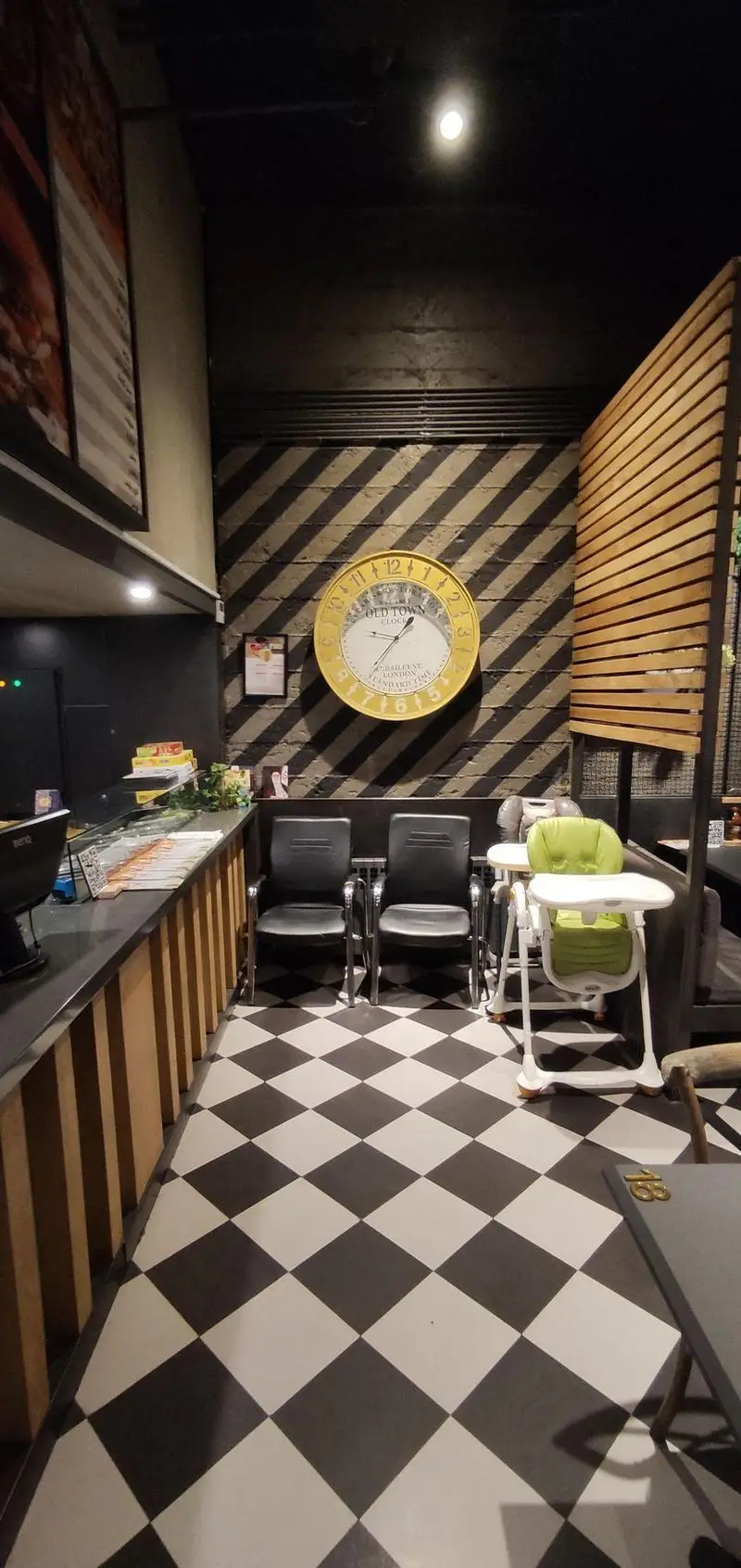 فضای داخلی رستوران ای تی اف شعبه کاشانی با طراحی مدرن شامل کفپوش شطرنجی، دیوار با خطوط مورب و ساعت بزرگ Old Town، به همراه صندلی‌های انتظار و صندلی کودک.