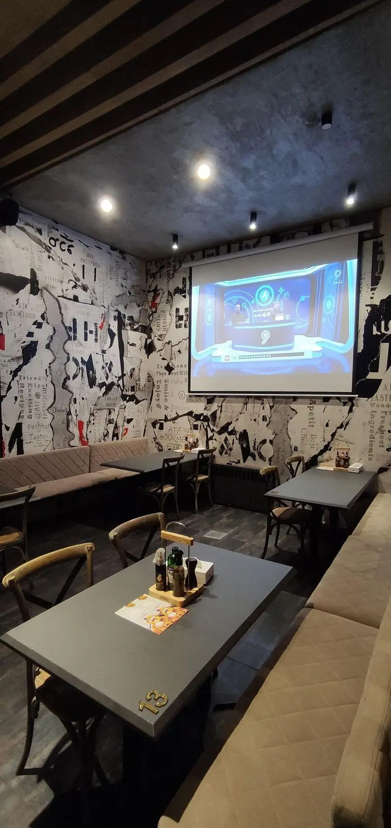 فضای داخلی مدرن و دنج رستوران ای تی اف شعبه کاشانی با دیوارهای طرح روزنامه، سقف بتنی، پروژکتور فعال و میز و صندلی‌های راحت.