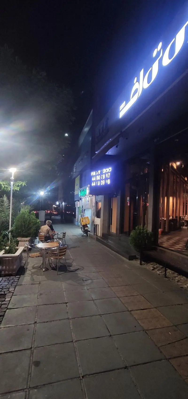 مشتریان در فضای باز فست فود ای تی اف شعبه کاشانی در شب، با تابلوی روشن و موتورسیکلت دلیوری آماده خدمت‌رسانی.