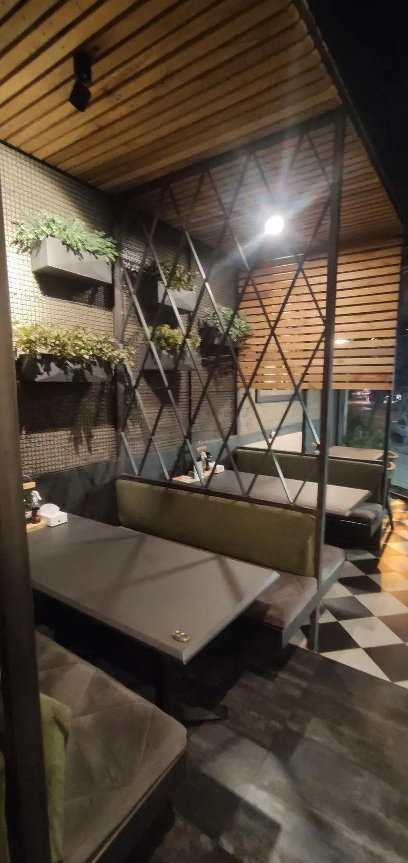 دکوراسیون داخلی کافه ای تی اف شعبه کاشانی: سقف چوبی، دیوارهای مشبک با گلدان‌های سبز، پارتیشن‌های فلزی مدرن، میز و صندلی‌های بوت شیک و نورپردازی گرم.