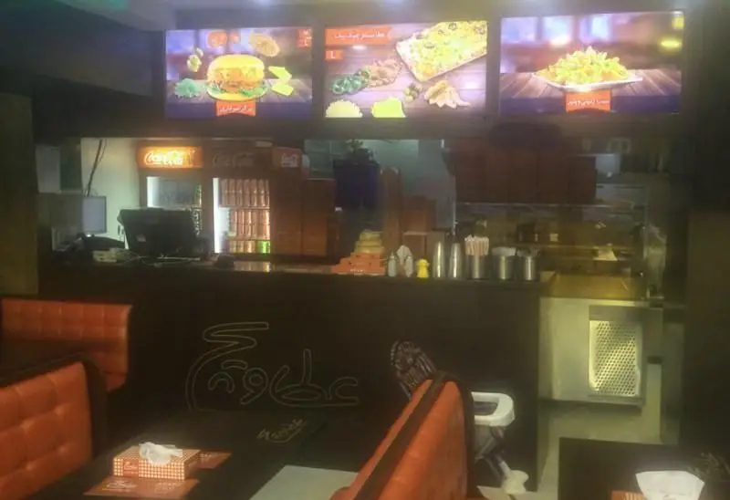 تصویر فضای داخلی رستوران عطاویچ اکباتان با منوی دیجیتال برگر، پیتزا، سیب‌زمینی سرخ‌کرده و نشیمن‌های نارنجی مشتریان.