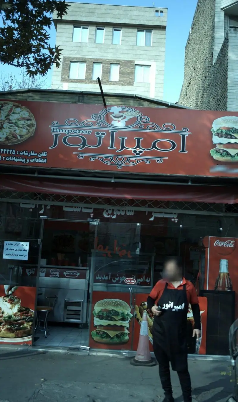 عکس فست فود امپراتور با تابلوی بزرگ شامل نام و لوگو، تصاویر پیتزا و همبرگر، و یکی از کارکنان با پیشبند امپراتور.