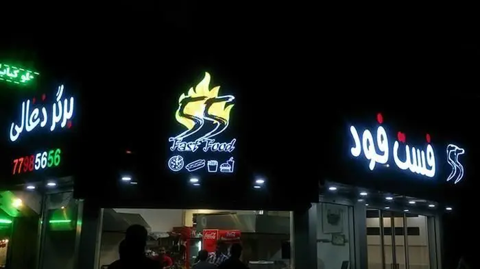 نمای خارجی فست فود ۵۵ در شب با تابلوهای درخشان نئونی، شامل نام "Fast Food 55" به فارسی و انگلیسی، لوگوی شعله‌ور و نمادهای غذاهای محبوب.
