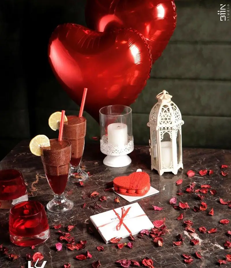 دکوراسیون رمانتیک کافه رستوران کالوک با بادکنک‌های قلبی، شمع، نوشیدنی‌های شیک و کیک ولنتاین روی میز مرمری با گلبرگ سرخ.