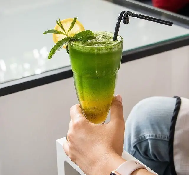دست در حال گرفتن لیوان بلند اسموتی خنک لایه‌ای سبز و زرد با تزئین نعنا و لیمو، یک نوشیدنی عالی در کافه رستوران کالوک.
