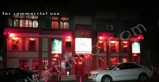 تصویر شبانه کسب و کار خانه کوچک، ساختمانی با نورپردازی قرمز و محیطی دلپذیر. خودروهای سفید و مشکی در جلو پارک شده‌اند.