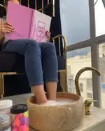 سالن زیبایی دنیز بیوتی: مشتری در حال ورق زدن کاتالوگ رنگ لاک ژل برای پدیکور. تجربه‌ای لوکس و آرامش‌بخش در کنار منظره شهر.