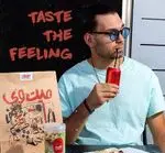مردی با ساندویچ و نوشیدنی کوکاکولا از غذای میت وی لذت می‌برد. بسته‌بندی جذاب و شعار "TASTE THE FEELING" در پس‌زمینه.