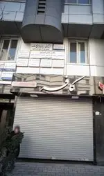 کافه با تابلوی برجسته سفید و کرکره بسته، همراه با تابلوهای مختلف پزشکان و آزمایشگاه در بالای ساختمان تجاری در شهر.