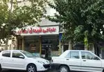 تصویری از نمای بیرونی مغازه طباخی شبهای تهران با تابلو قرمز رنگ، در خیابانی پردرخت با دو خودروی سفید پارک شده در جلو