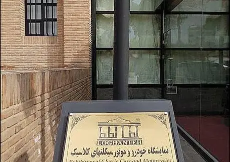تصویری از نمای ورودی کافه لقانطه و تابلوی نمایشگاه خودرو و موتورسیکلت‌های کلاسیک در ساختمانی با معماری آجری و شیشه‌ای مدرن.