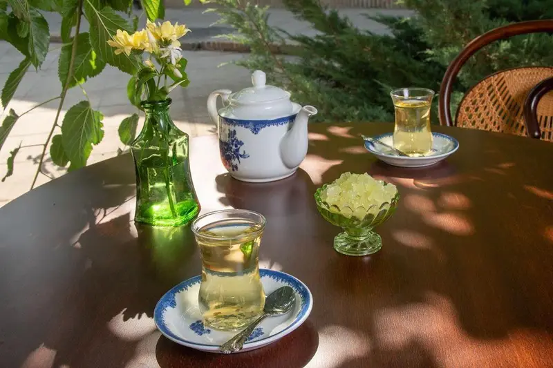 میز زیبای چای سنتی کافه لقانطه با گلدان گل و قوری سفید، به همراه چای و نبات در فضای دلنشین و آرامش‌بخش، مناسب استراحت.