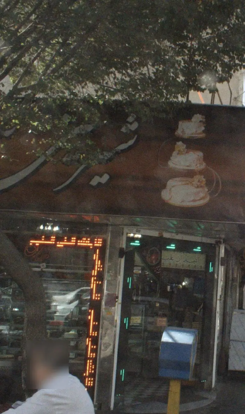 تصویر نمای بیرونی مغازه شیرینی تبریزی، تابلوی بزرگ با طرح کیک و انواع شیرینی و دسر، با ویترین نورانی و درخت