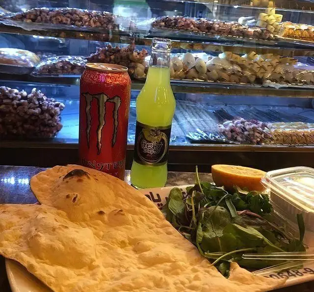 سینی غذای کامل در جگر ذغالی پامچال: نان تازه، سالاد سبز و نوشیدنی‌های متنوع با ویترین پر از شیرینی و کباب در پس‌زمینه.