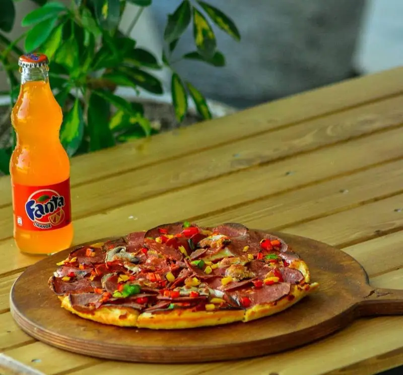 لذت طعم پیتزا پرملات فست فود کاوان با پپرونی، فلفل دلمه‌ای، قارچ و نوشابه خنک روی میز چوبی برای یک وعده عالی.