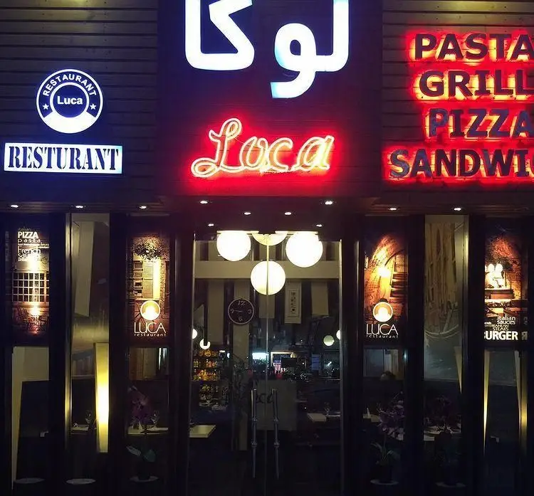 تصویر جلوی کافه رستوران لوکا در شب با نورپردازی نئونی روشن، شامل نام لوکا و منوی غذاهای پیتزا، پاستا، گریل و ساندویچ.