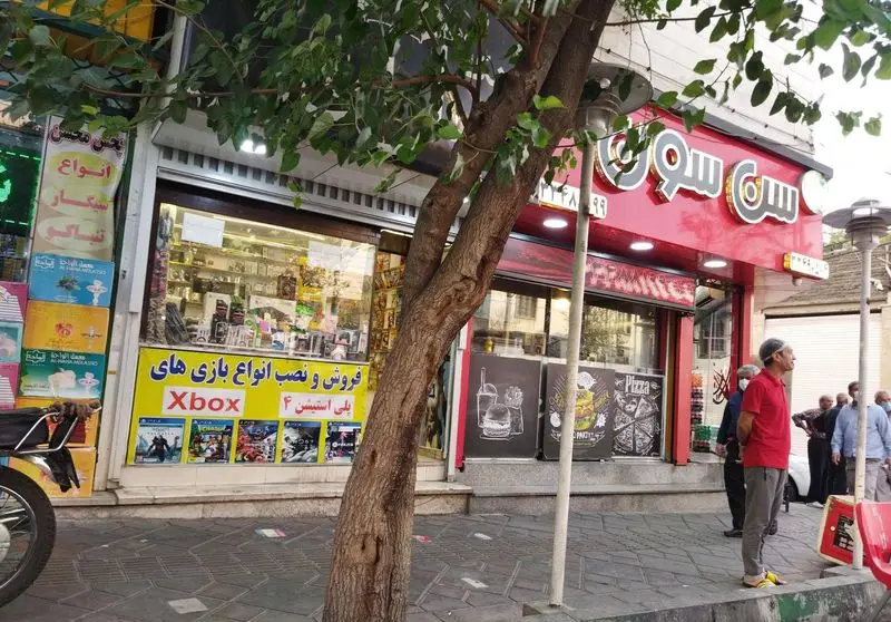 نمای بیرونی فست فود ساندویچ سن سون با تابلوی قرمز و مغازه بازی‌های ایکس باکس و پلی استیشن ۴ در کنارش، در یک روز آفتابی.