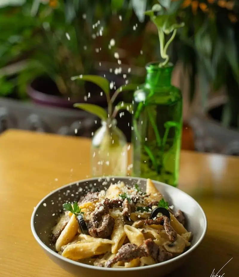 پاستای لذیذ با گوشت و قارچ در کافه لیو، همراه با پنیر رنده شده و تزئینات سبز. غذایی ایتالیایی در فضایی دلنشین و سرسبز.