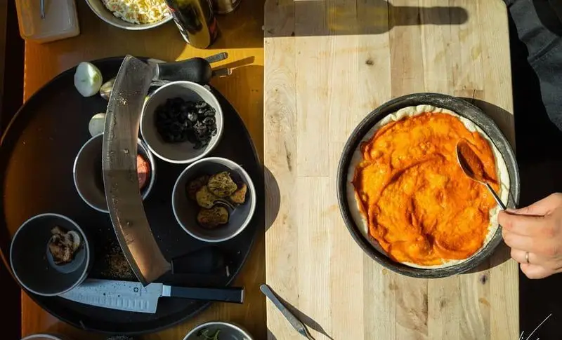 مراحل تهیه پیتزای کافه لیو: پخش سس مخصوص نارنجی روی خمیر تازه، همراه با مواد اولیه متنوع شامل زیتون و مرغ برای یک تجربه دلپذیر.