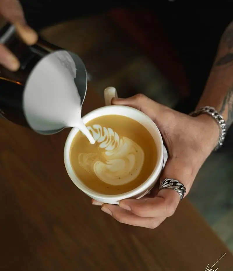 دست‌های باریستا در کافه لیو با مهارت شیر را برای خلق لاته آرت شگفت‌انگیز در فنجان قهوه می‌ریزند. با دستبند و انگشتر زیبا.