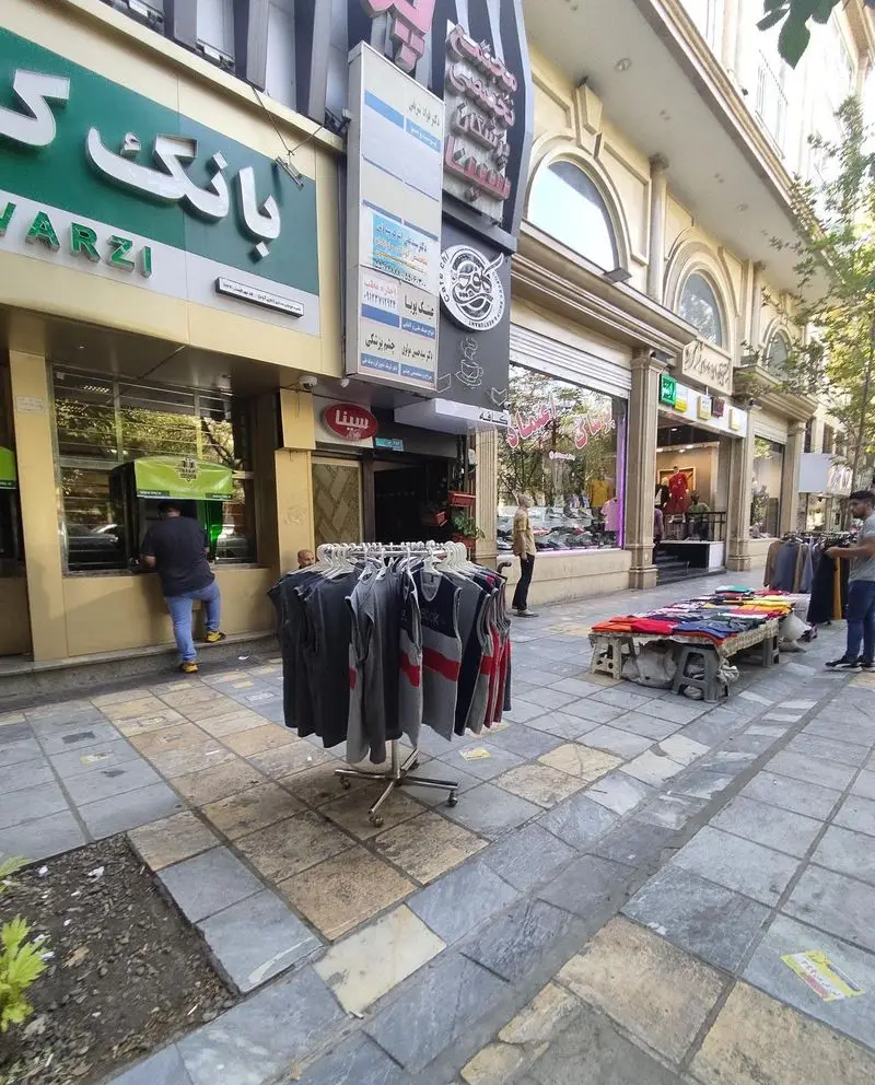 چايخانه رهروان در خیابانی شلوغ با فروشگاه‌های لباس، بانک و عابربانک در حال ارائه خدمات به مشتریان در یک روز آفتابی.