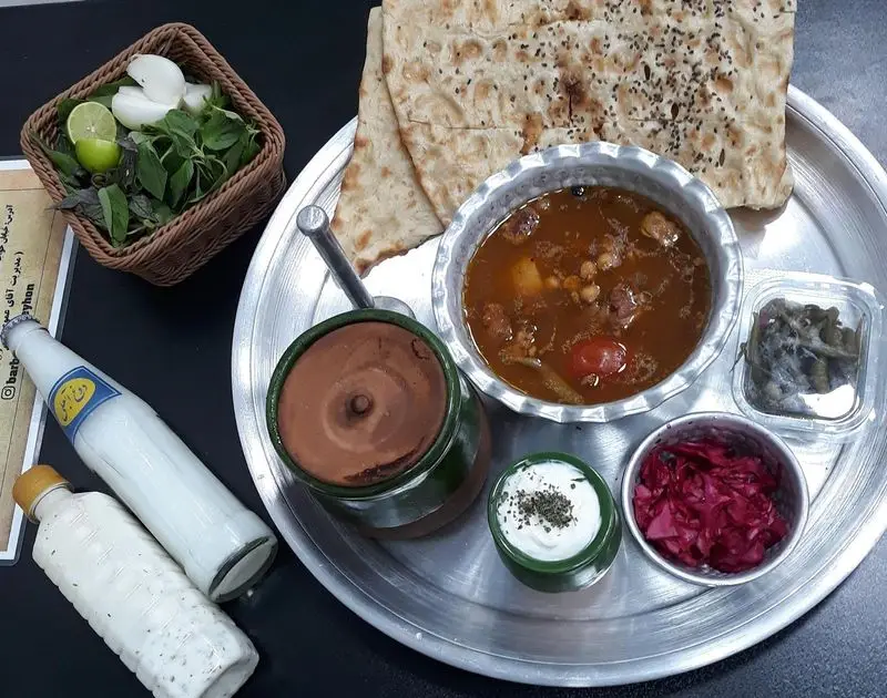 تصویری از میز غذای سنتی ایرانی در باربیکیو ریحون: آبگوشت دیزی با نان سنگک، دوغ، سبزی خوردن، ماست و ترشی لذیذ.