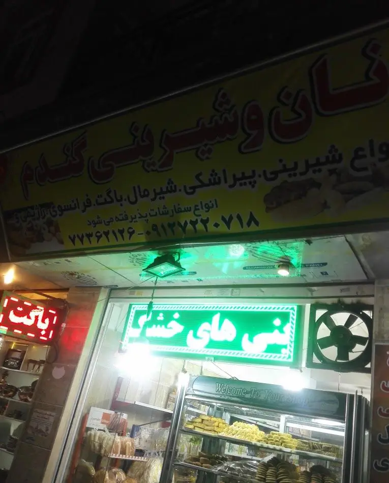 نمای بیرونی مغازه نان و شیرینی گندم با تابلو بزرگ زردرنگ، نمایش انواع شیرینی، نان، پیراشکی، باگت و شماره تماس برای سفارشات.