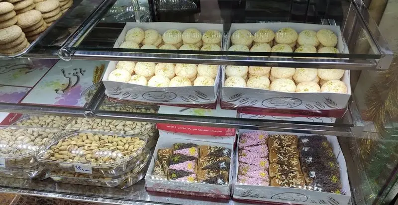 ویترین قنادی نان و شیرینی گندم با انواع شیرینی‌های سنتی گرد، نان‌های گندمی، کیک‌های رنگی و پسته تازه برای مشتریان