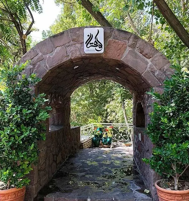 تصویری از ورودی رستوران کوهستان با لوگوی مجموعه بر فراز طاق سنگی، گلدان‌های سرسبز و مسیری زیبا در دل طبیعت.