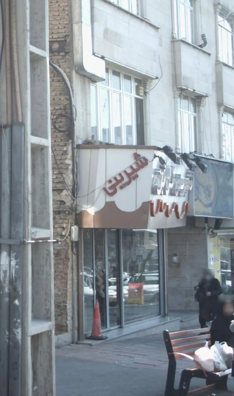 تصویری از نمای خارجی قنادی شیرینی گل بن، با تابلوی قرمز رنگ "شیرینی" و تزئینات شکلاتی، کنار پیاده‌رو شهری. ساختمان روشن.