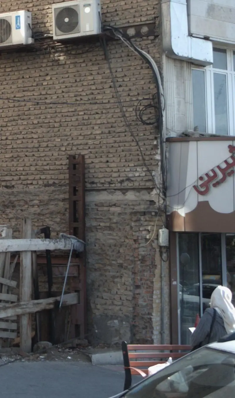 تصویری از نمای خارجی شیرینی گل بن: ساختمان آجری با دو کولر گازی، سیم‌کشی برق و تابلوی فروشگاه، در کنار نیمکت مشتریان.