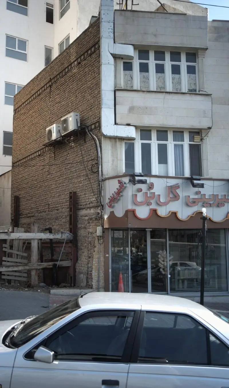 شیرینی گل بن: نمای خیابانی مغازه با تابلوی درخشان، پنجره‌های بزرگ و سبک معماری منحصربه‌فرد، منعکس‌کننده فضای شهری.