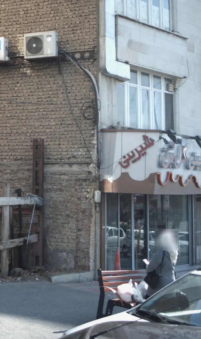 نمای بیرونی قنادی شیرینی گل بن با تابلوی قرمز "شیرینی"، شامل بخش‌هایی از ساختمان آجری و پیاده‌رو پرتردد.