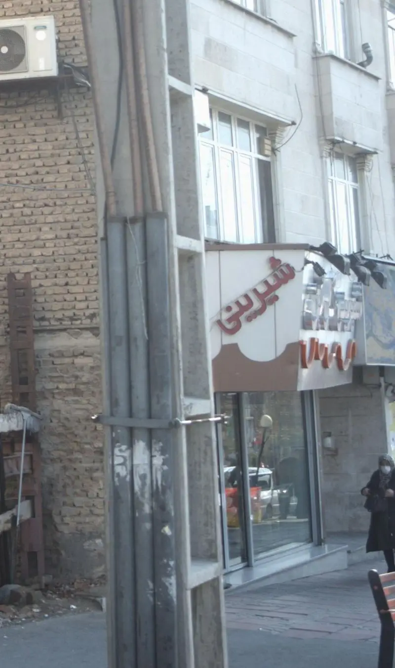تصویری از نمای خیابان شیرینی گل بن، با تابلوی روشن "شیرینی" بر سردر، ساختمان سنگی و عابری با ماسک در پیاده‌رو شهر.
