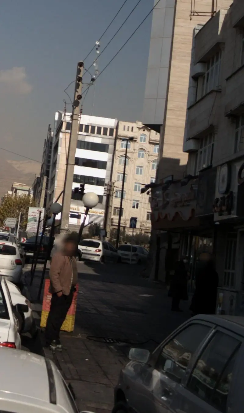 تصویری از نمای خارجی قنادی شیرینی گل بن در یک خیابان شلوغ شهری، با مردم، ساختمان‌های بلند و خودروها در روز آفتابی.
