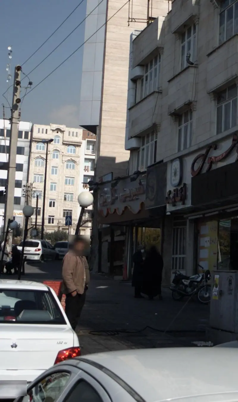 تصویری از نمای بیرونی قنادی شیرینی گل بن در یک روز آفتابی، با ساختمان‌های بلند اطراف، عابران پیاده و ترافیک شهری.