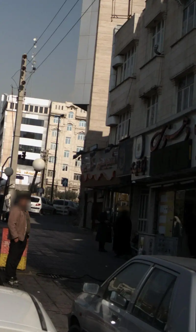 نمای شهری از شیرینی گل بن: خیابان شلوغ با ساختمان‌های متنوع، مردم و خودروها در روز آفتابی.