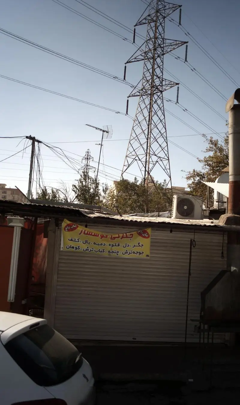 نمای بیرونی جگرکی کوهسار در یک روز آفتابی با تابلوی زرد حاوی نام و انواع کباب‌ها (جگر، دل، قلوه، جوجه ترش، چنجه). برج برق و سیم‌های معلق دیده می‌شود.