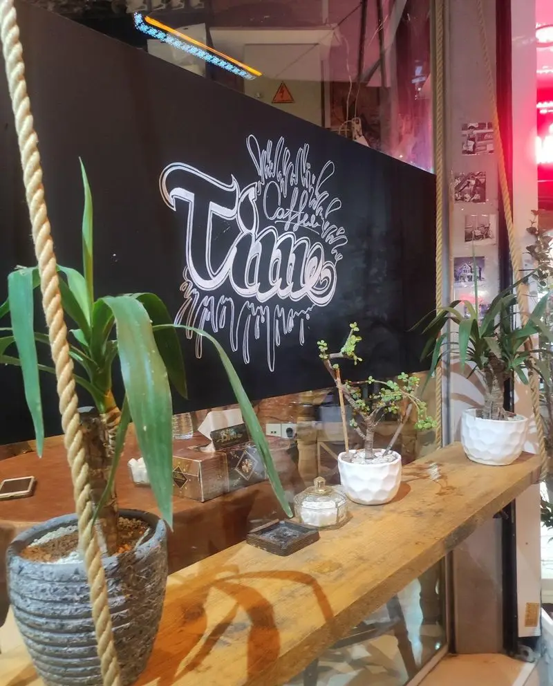 تصویری از فضای دلنشین کافه تایم: تابلوی "قهوه و آبمیوه" بر دیوار، گیاهان طبیعی و قفسه‌ای روستیک با طناب‌های کنفی.