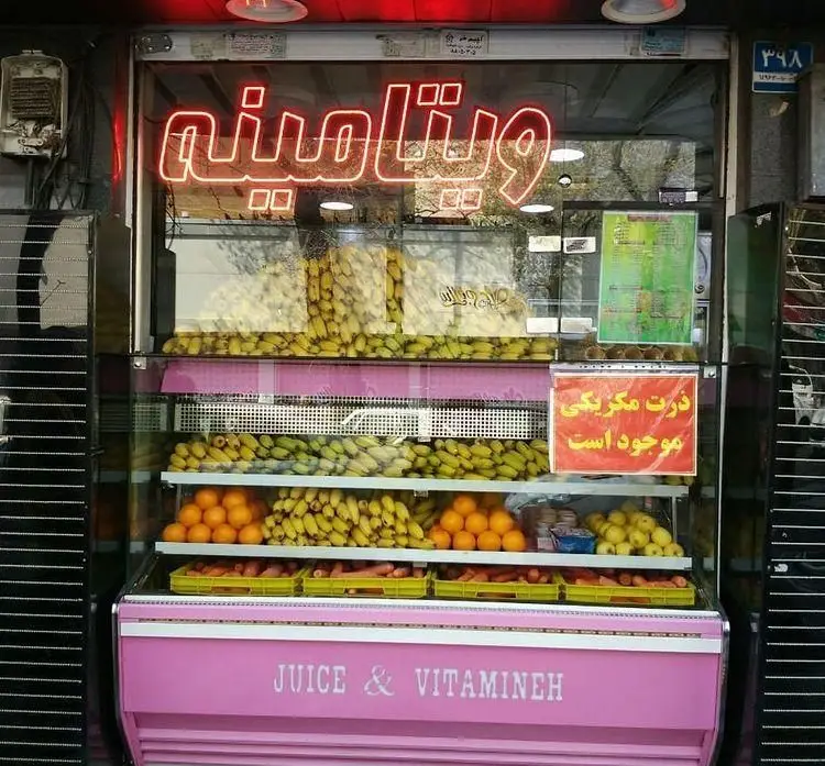 ویترین کافه بستنی صمد، فروشگاه آبمیوه و ویتامینه با نئون روشن "ویتامینه"، میوه‌های تازه، موز، پرتقال و ذرت مکزیکی.