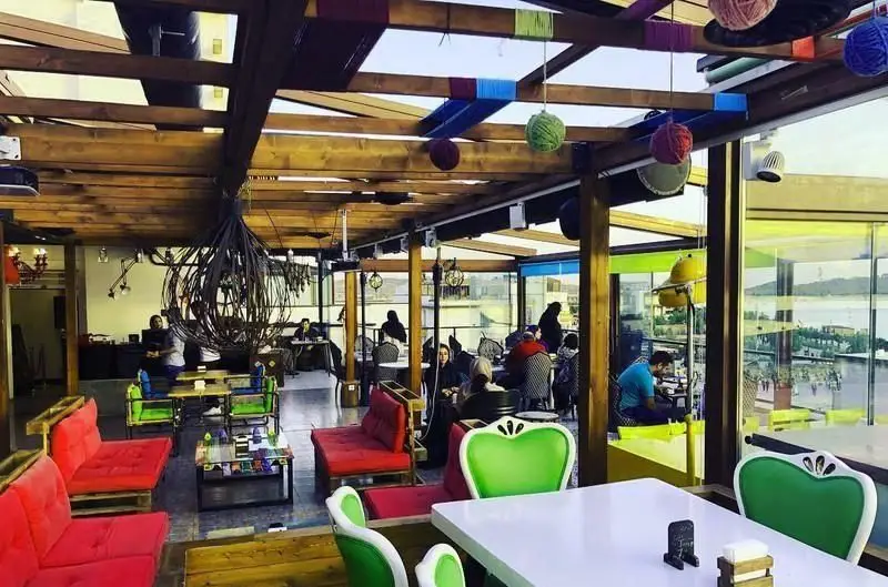 فضای داخلی کافه رستوران آنتیکا با سقف چوبی، تزئینات رنگی خلاقانه، مبلمان قرمز و سبز، و پنجره‌های وسیع مشرف به منظره آب.