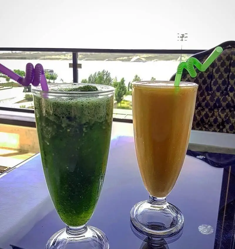 دو نوشیدنی تازه و گوارا، اسموتی سبز و آبمیوه پرتقالی، در کافه رستوران آنتیکا با چشم‌اندازی زیبا و فضایی دلنشین برای مشتریان.