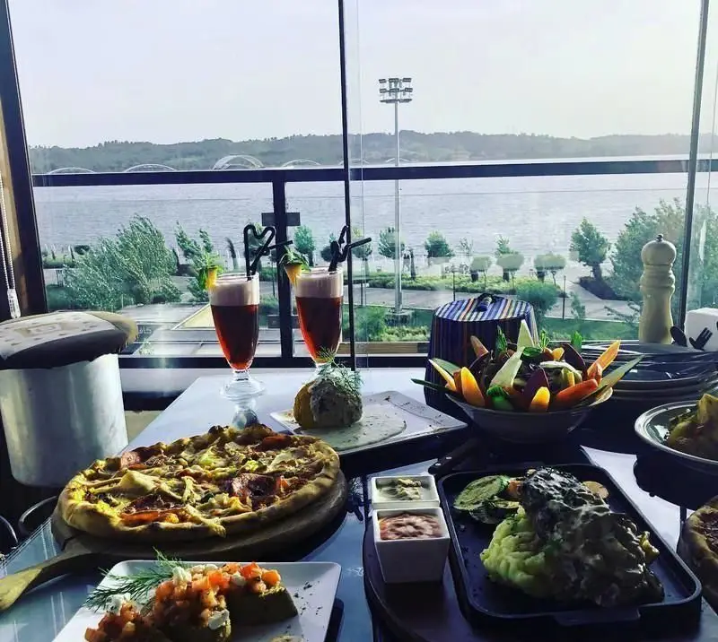 کافه رستوران آنتیکا: میزی پر از غذاهای لذیذ شامل پیتزا، سالاد، غذای اصلی و نوشیدنی با چشم‌انداز خیره‌کننده دریاچه و محیطی آرامش‌بخش.