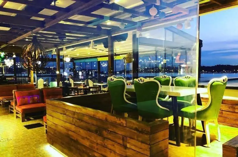 فضای داخلی دلنشین کافه رستوران آنتیکا با میز و صندلی‌های سبز لوکس و مبلمان چوبی-قرمز، رو به منظره تماشایی دریا و چراغ‌های شهر در غروب.