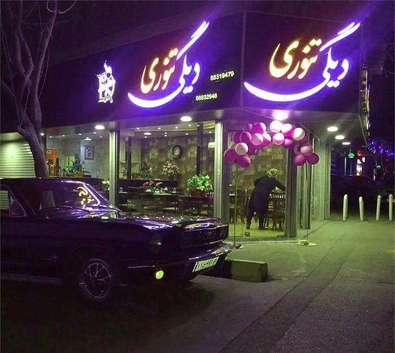 نمای شب رستوران ایرانی دیگی تنوری با نورپردازی بنفش، موستانگ کلاسیک و بادکنک‌های رنگی، فضایی جذاب برای سرو غذای سنتی ایرانی.