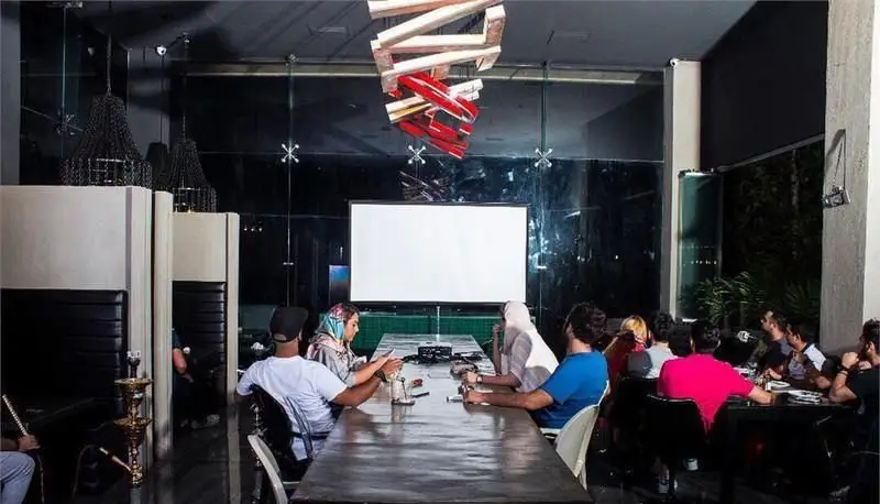 فضای داخلی کافی شاپ راندوو با افراد دور میز در حال گفتگو و پرده نمایش. محیطی مدرن برای جلسات و دورهمی دوستانه.