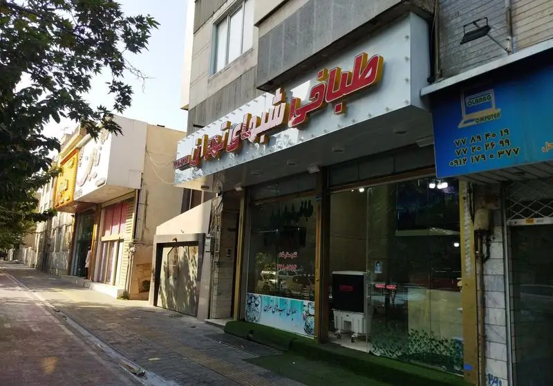 تصویری از مغازه طباخی شبهای تهران، یک رستوران سنتی در خیابان‌های تهران، با نمای بیرونی روشن، برگ‌های سبز و ساختمان‌های شهری.